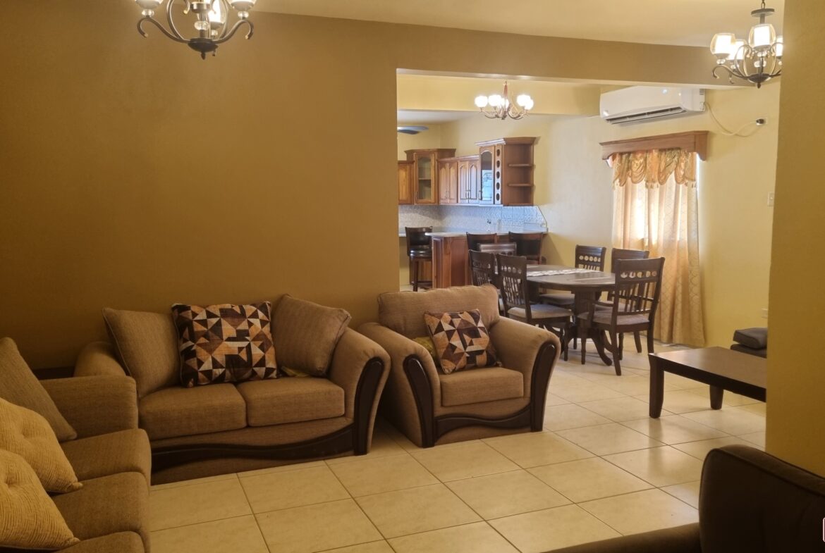 Open concept living room area leading into kitchen in townhouse for sale. The living room has a couch, coffee table, fireplace, and a large window. The kitchen has stainless steel appliances, granite countertops, and a breakfast bar.