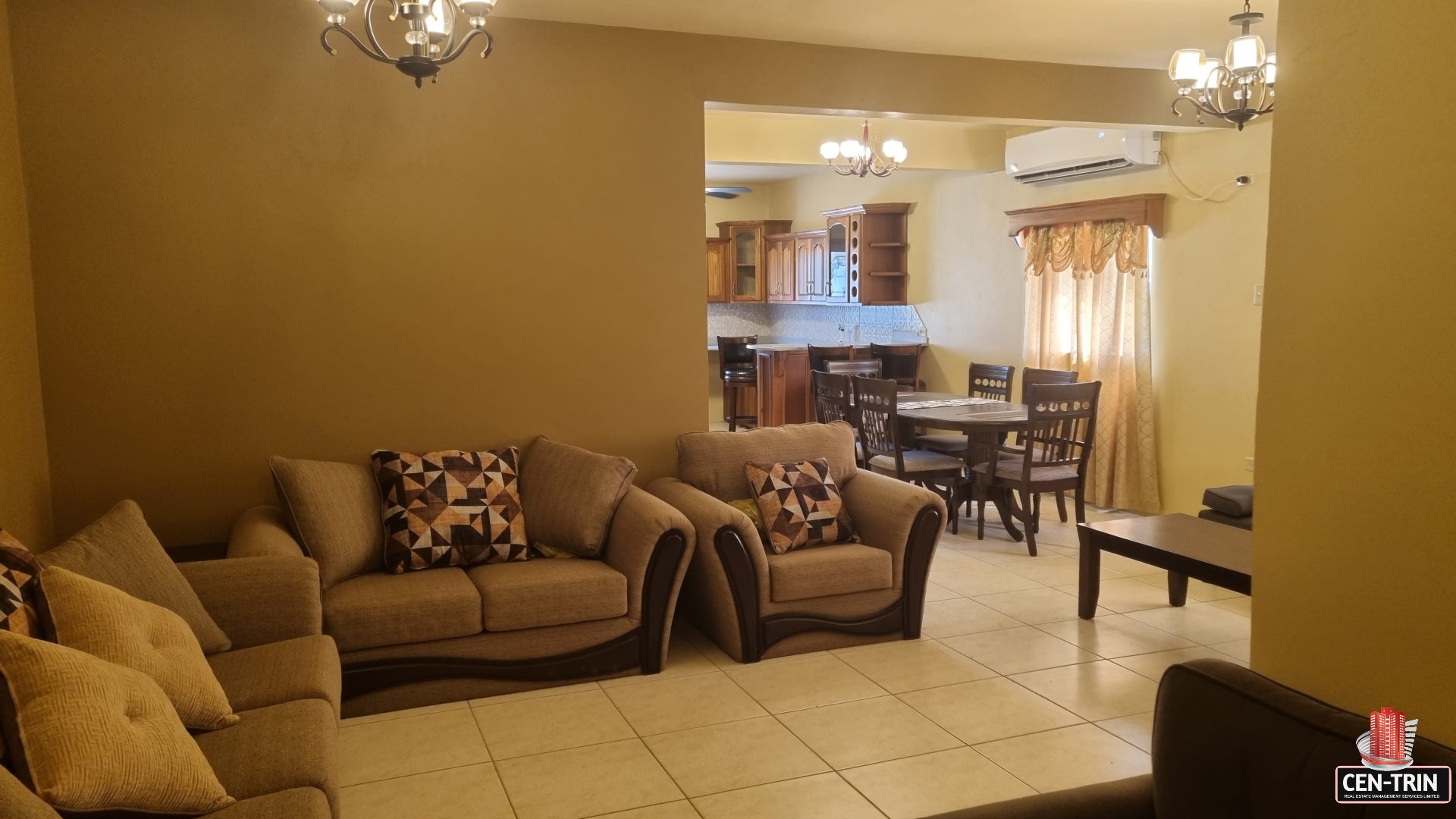 Open concept living room area leading into kitchen in townhouse for sale. The living room has a couch, coffee table, fireplace, and a large window. The kitchen has stainless steel appliances, granite countertops, and a breakfast bar.