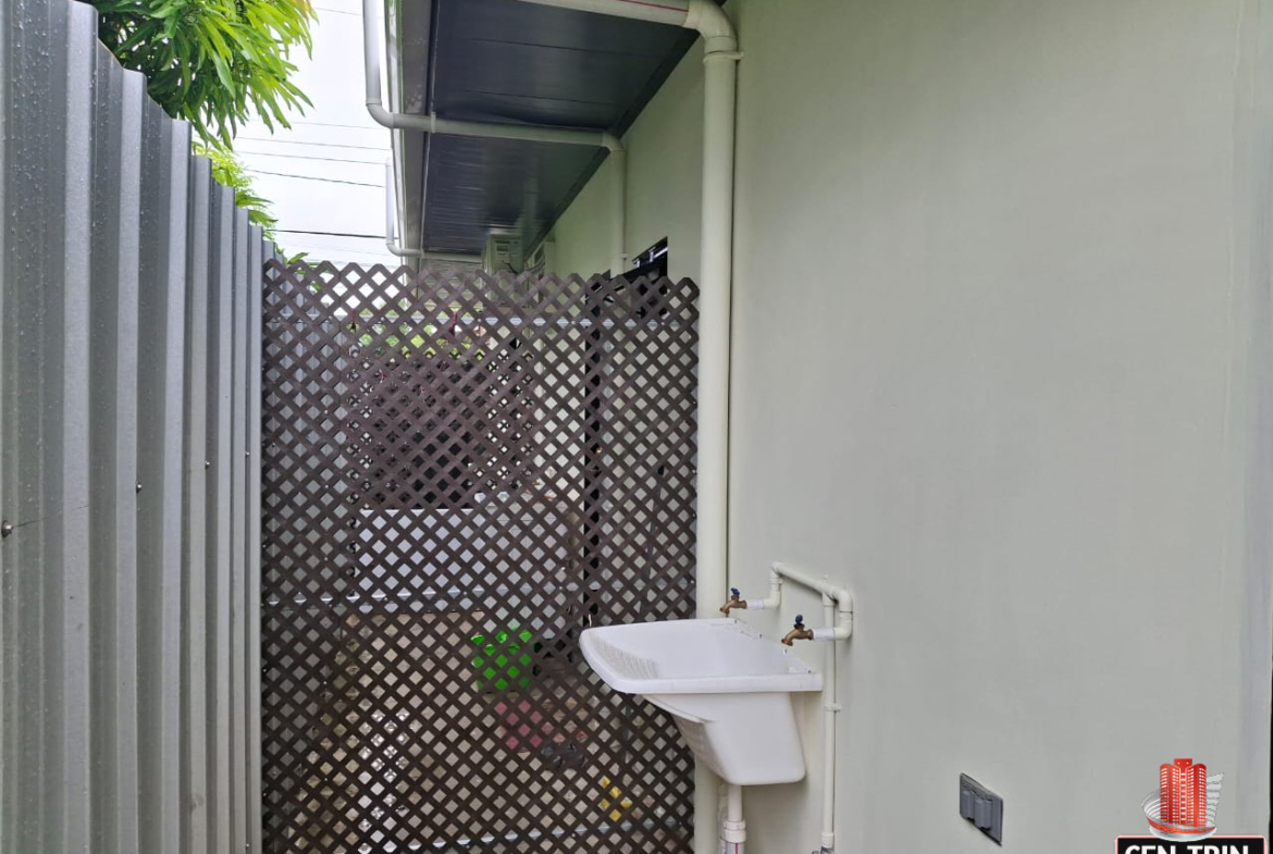 Exterior view of a section of an apartment building with light green walls, showing a white utility sink mounted on the wall, white PVC pipes, a gray metal fence, and a brown lattice fence. Green foliage is visible at the top.