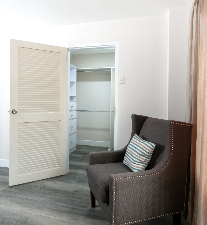 Master bedroom at Roystonia Mews featuring a wing chair, open walk-in closet with built-in shelving, and gray plank flooring.