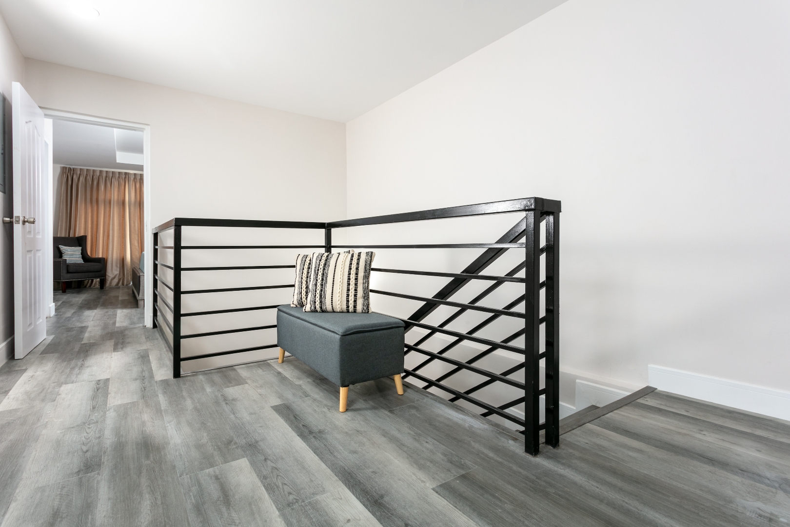 Upstairs hallway at Roystonia Mews featuring a modern black railing, gray plank flooring, and a small bench with a striped cushion.