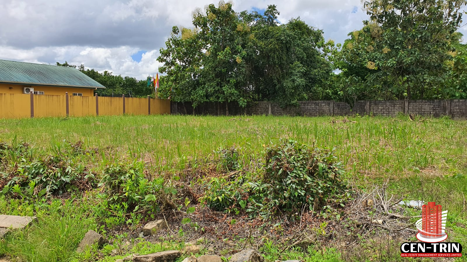 A wide view of the two combined plots of land for sale in Trinidad, showing lush green grass, some overgrowth, and surrounding fences/buildings, indicating a residential setting in Krystal Gardens.