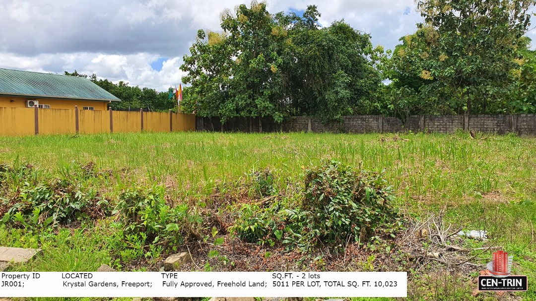 A wide view of the two combined plots of land for sale in Trinidad, showing lush green grass, some overgrowth, and surrounding fences/buildings, indicating a residential setting in Krystal Gardens.