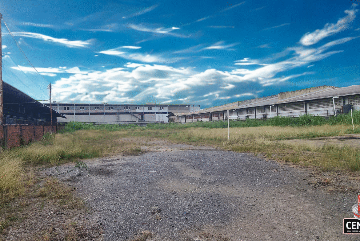 Ground-level view of the commercial land for rent on Patraj Trace, Aranguez, showing a gravel and dirt foreground leading to an open grassy area, with commercial buildings in the background under a dramatic blue sky.
