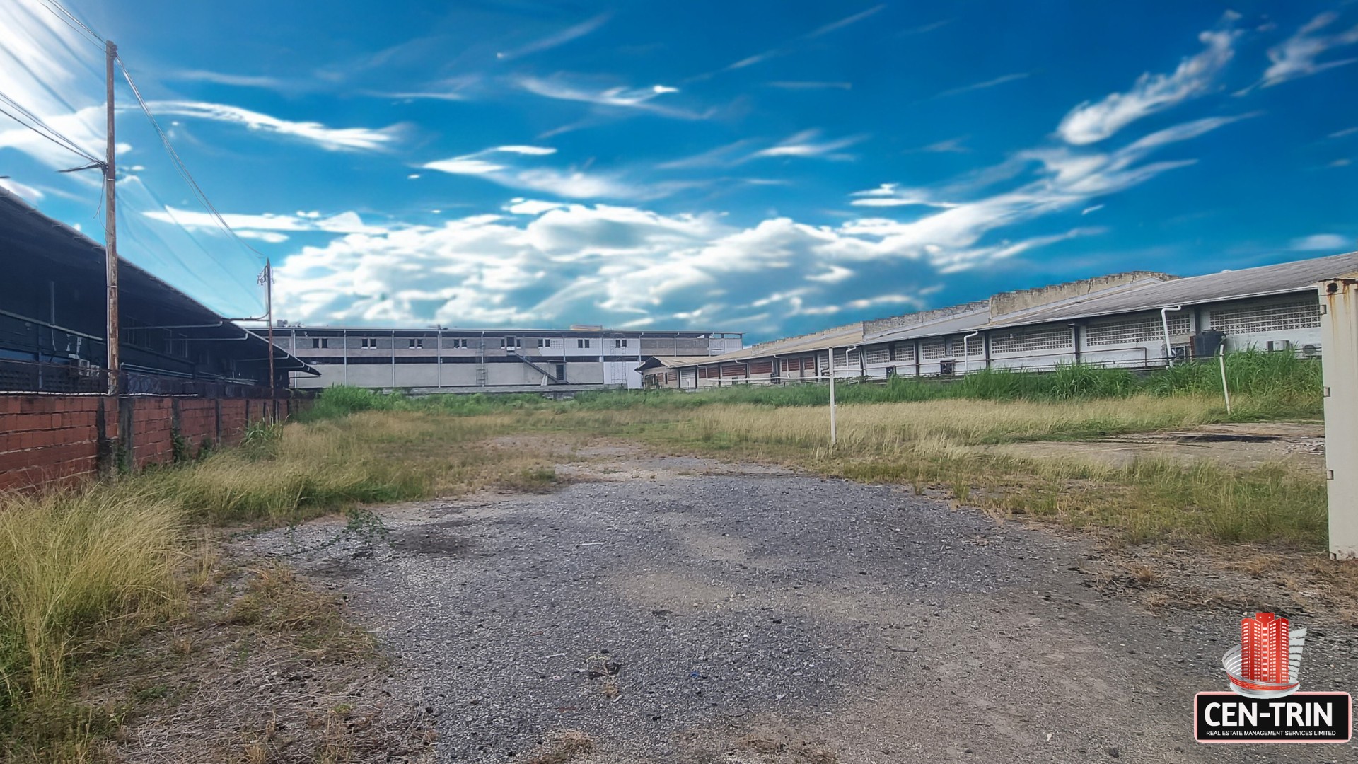 Ground-level view of the commercial land for rent on Patraj Trace, Aranguez, showing a gravel and dirt foreground leading to an open grassy area, with commercial buildings in the background under a dramatic blue sky.