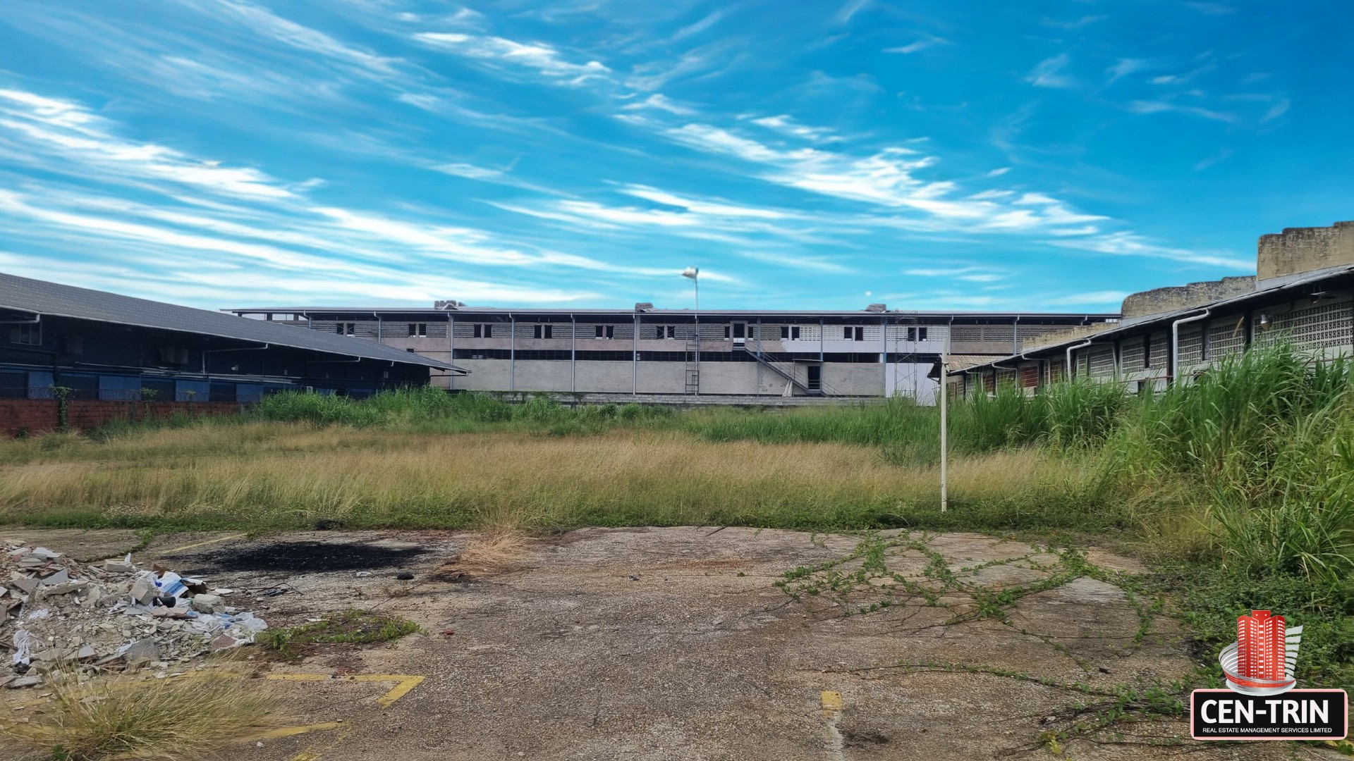 Ground-level view of the commercial land for rent on Patraj Trace, Aranguez, showing a partially cleared foreground with debris, leading to an open grassy area, bordered by large industrial buildings under a dynamic sky.