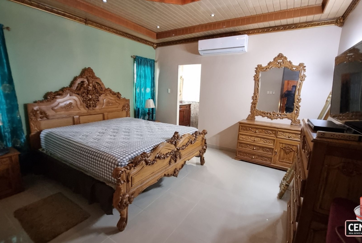 Full view of the beautifully furnished bedroom with carved wooden bed, dresser, air conditioning, and direct en-suite access in this luxury home for sale in Cunupia.
