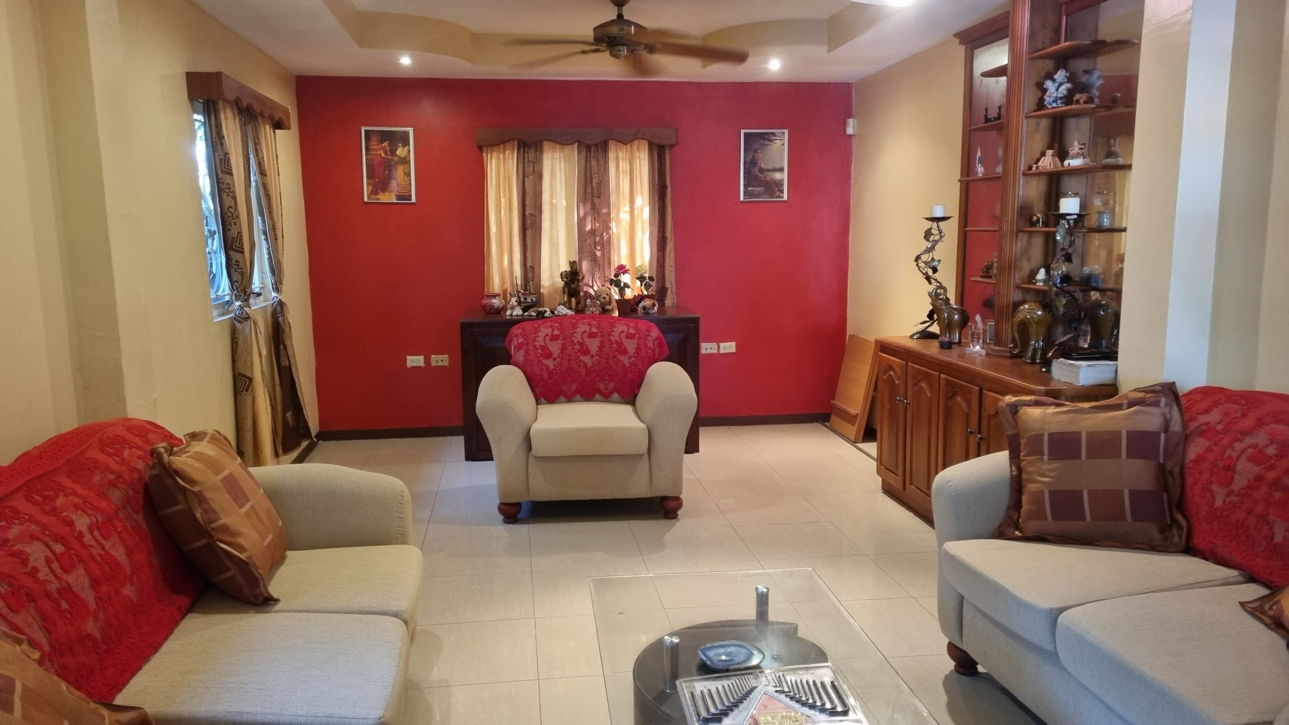Spacious Living Room in Fully Furnished House for Sale Interior view of a living room in a fully furnished house for sale, featuring red accent walls, two light-colored sofas with red throws, a single armchair, and a wooden display cabinet.
