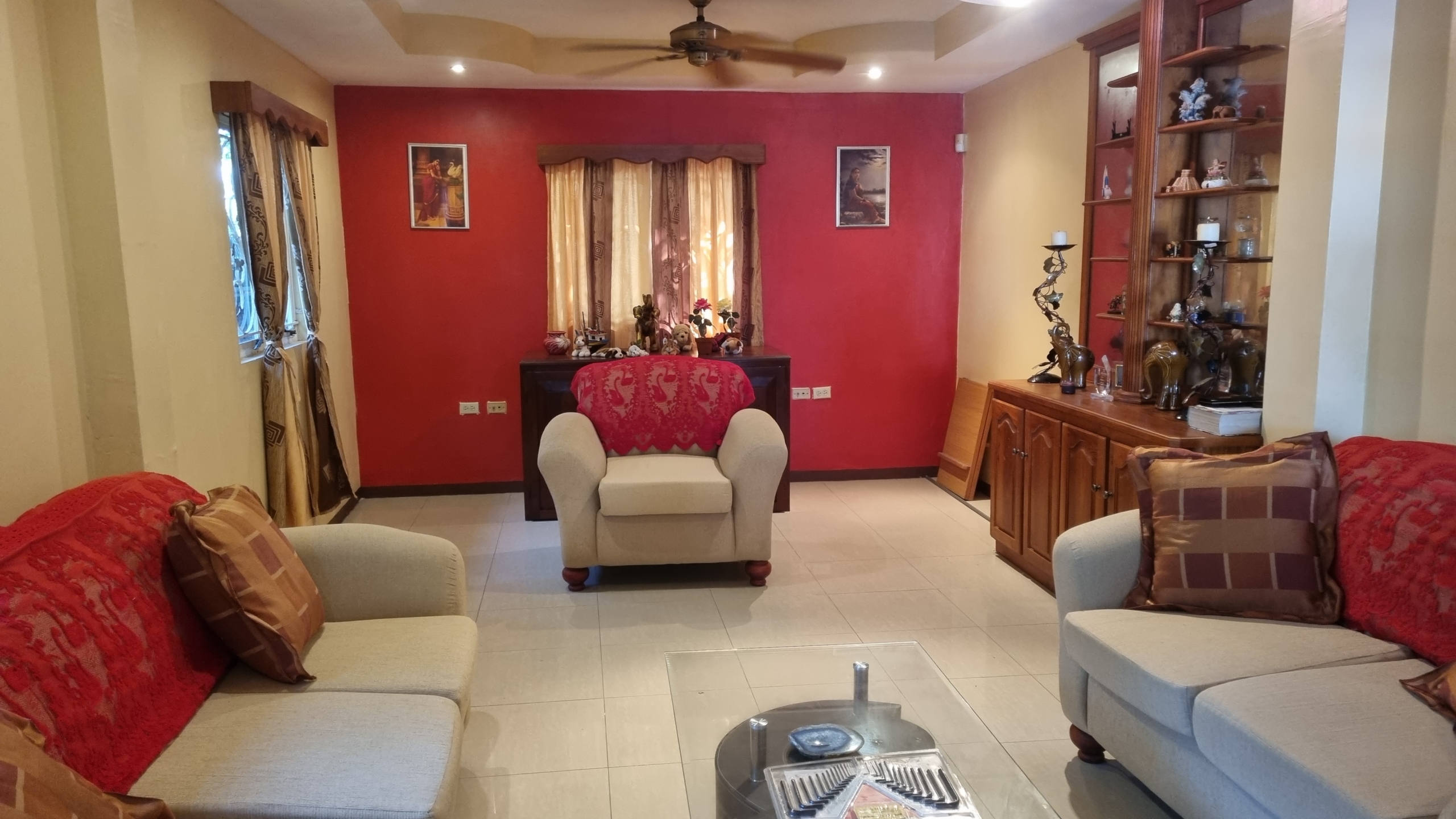 Interior view of a living room in a fully furnished house for sale, featuring red accent walls, two light-colored sofas with red throws, a single armchair, and a wooden display cabinet.