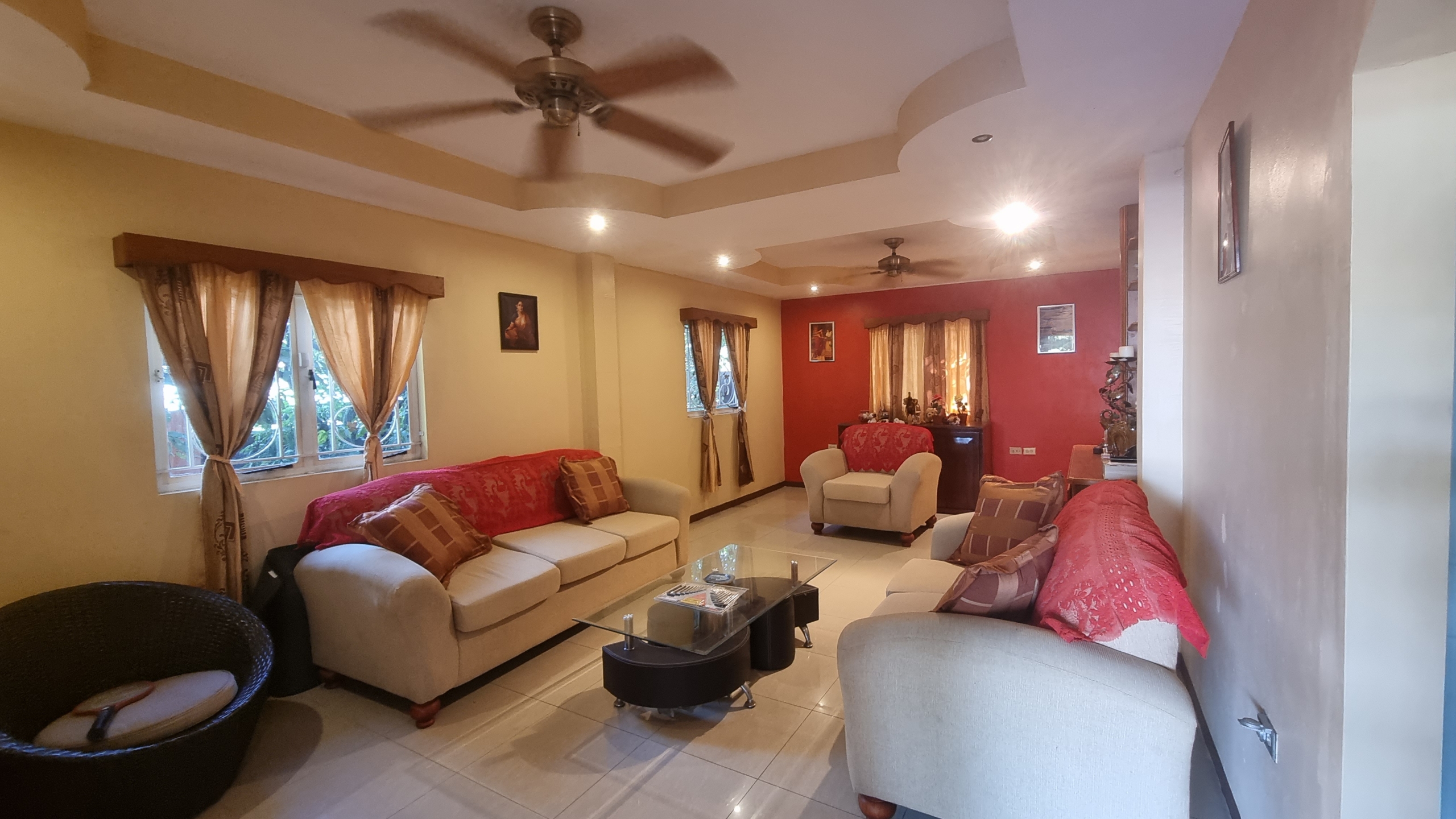 Wide view of a spacious living room and family area in a fully furnished house for sale, showing multiple seating arrangements, ceiling fans, and red accent walls.