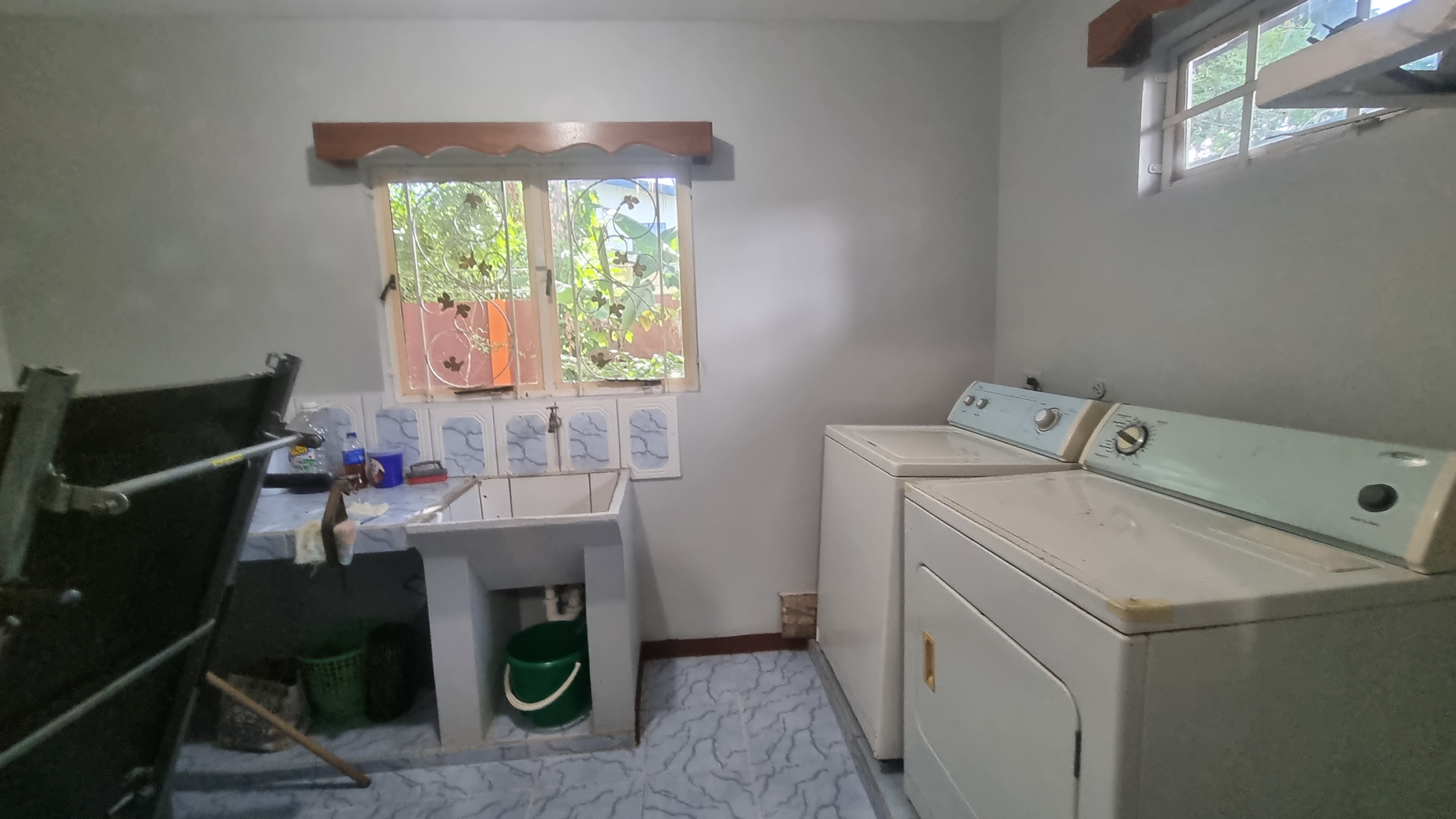 A functional laundry room in a fully furnished house for sale, with a washing machine, dryer, utility sink, and window overlooking a green outdoor area.