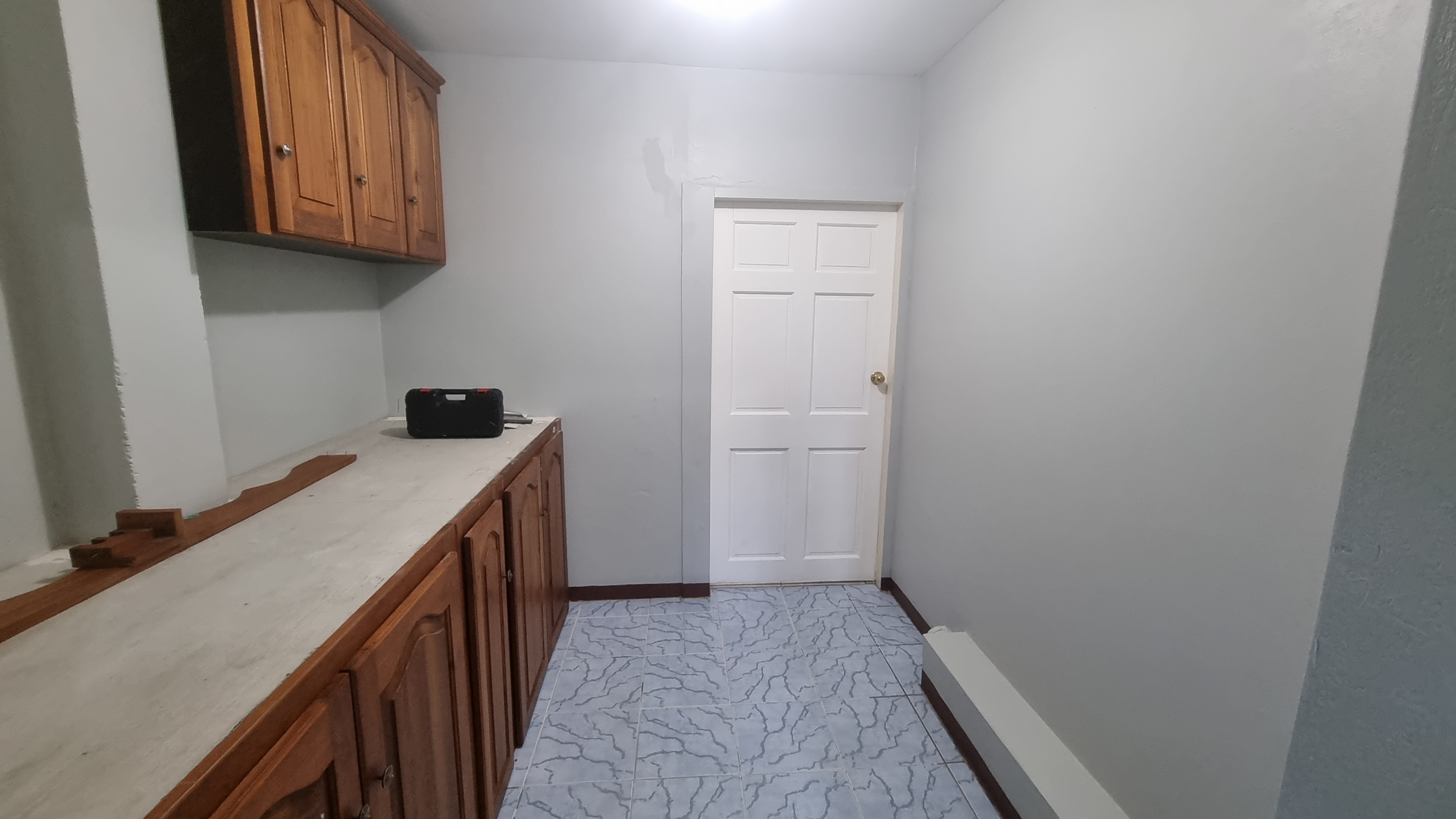 A hallway or utility space in a fully furnished house for sale, featuring wooden cabinets, light-colored walls, tiled floor, and a white door leading to another room.