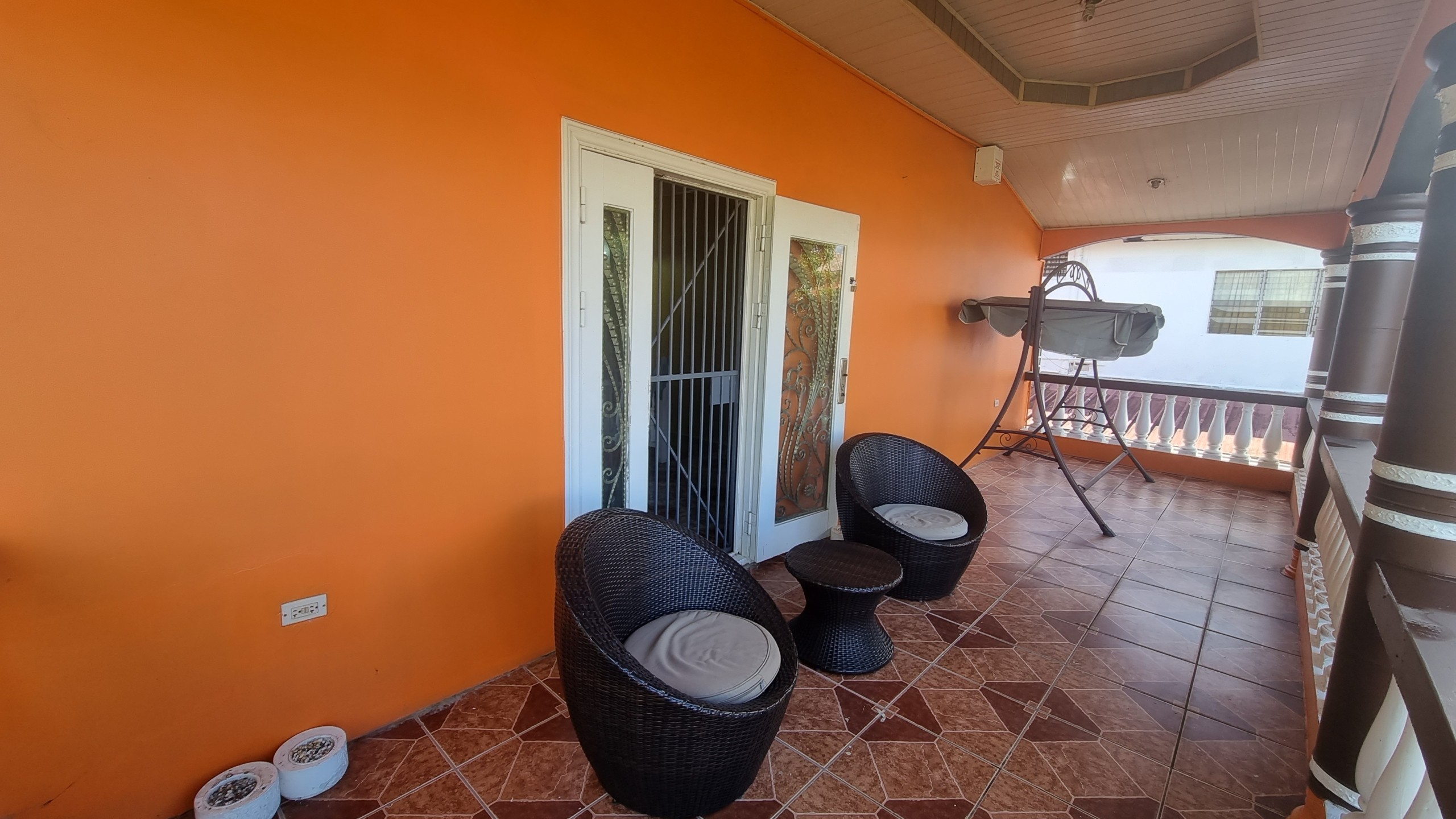 Inviting Balcony Space in Fully Furnished House for Sale A cozy balcony area of a fully furnished house for sale, with outdoor seating, an orange wall, tiled floor, and a view of the surrounding neighborhood.
