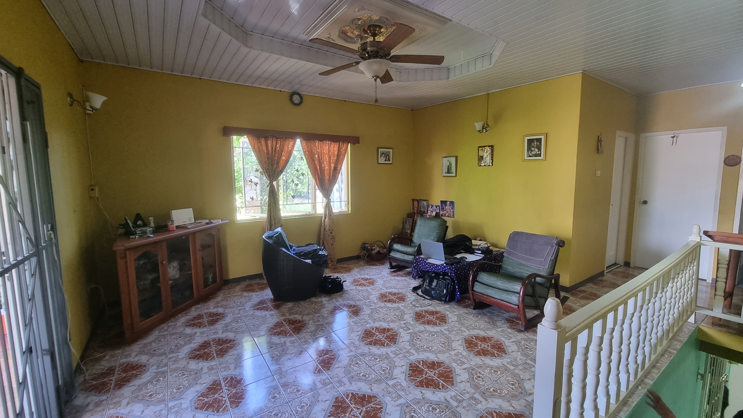 A first-floor family room or den in a fully furnished house for sale, with yellow walls, tiled floor, seating, and a view of the staircase.