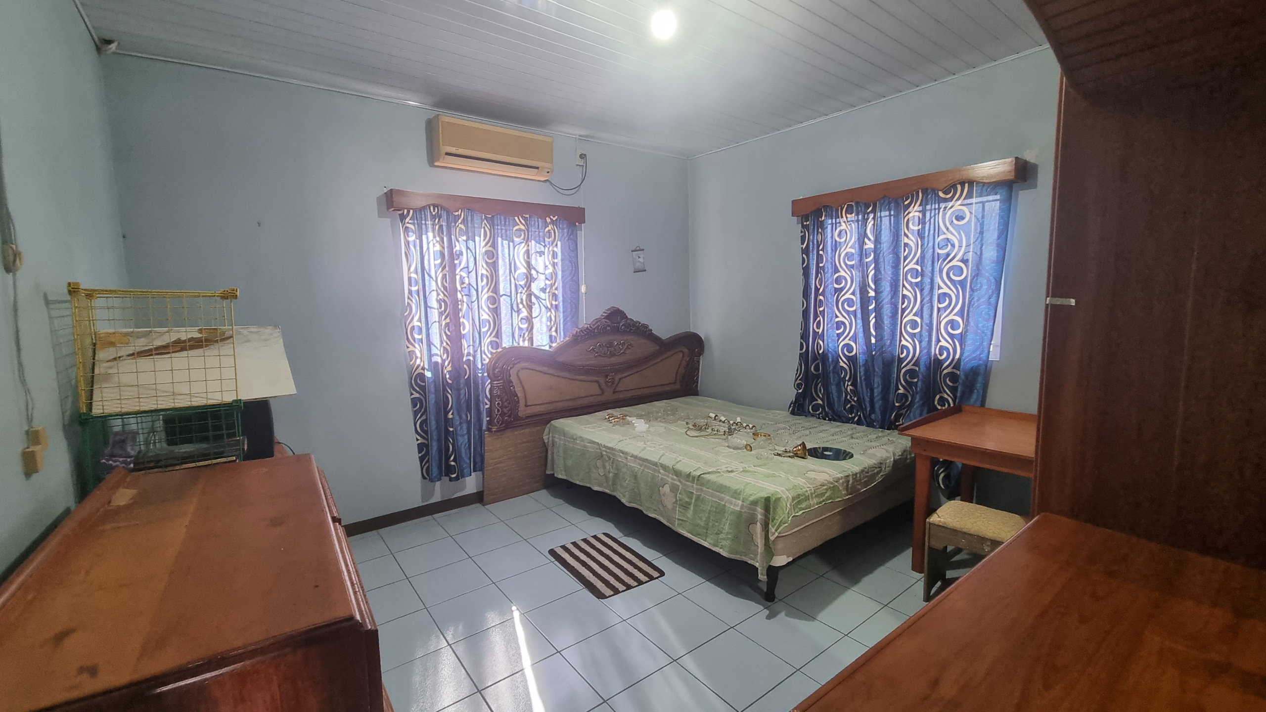 A bedroom in a fully furnished house for sale, featuring a large bed with an ornate headboard, an air conditioning unit, wooden furniture, and blue patterned curtains.