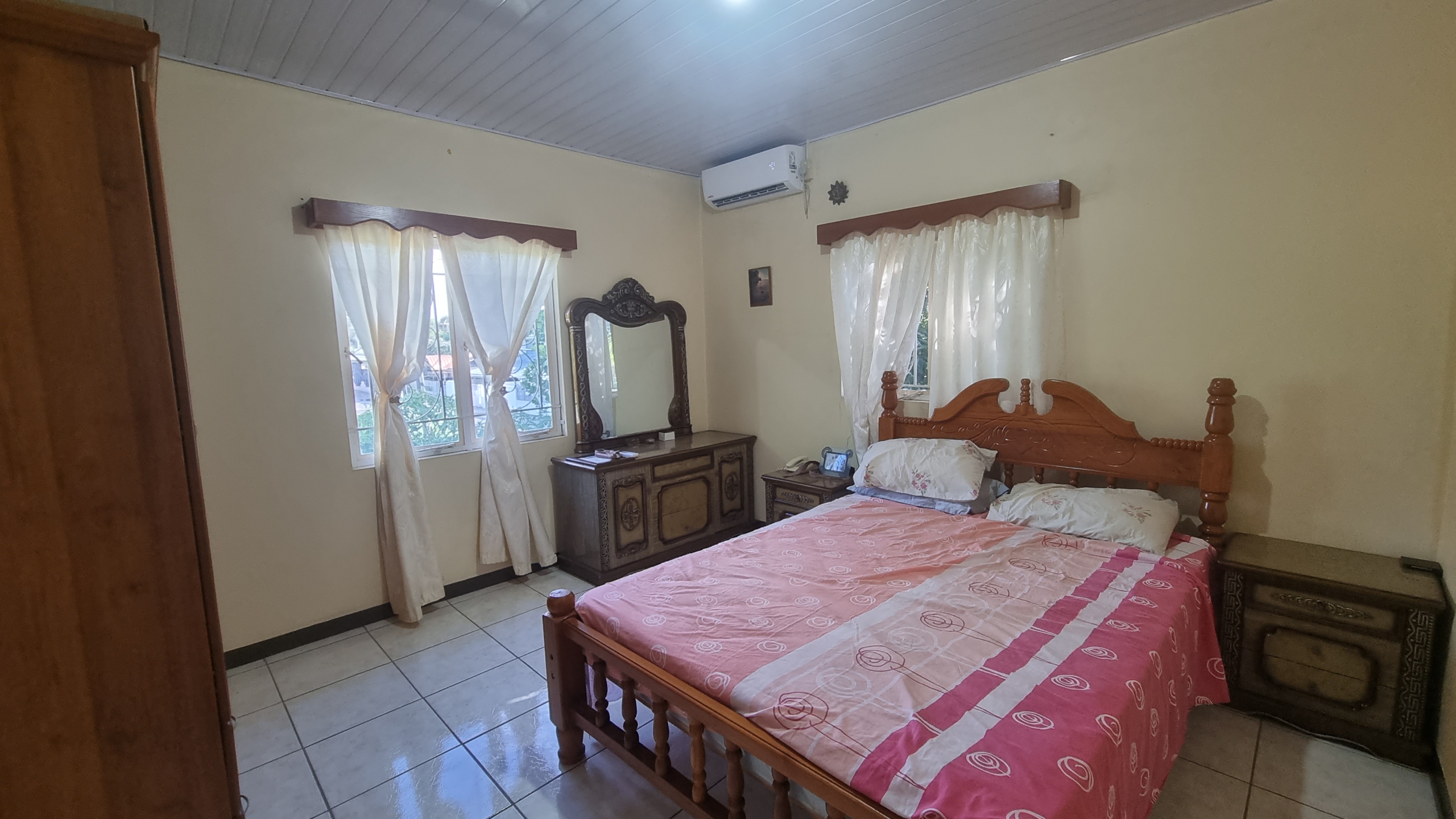 Another comfortable bedroom in a fully furnished house for sale, featuring a wooden bed frame, dresser with mirror, nightstands, and air conditioning.