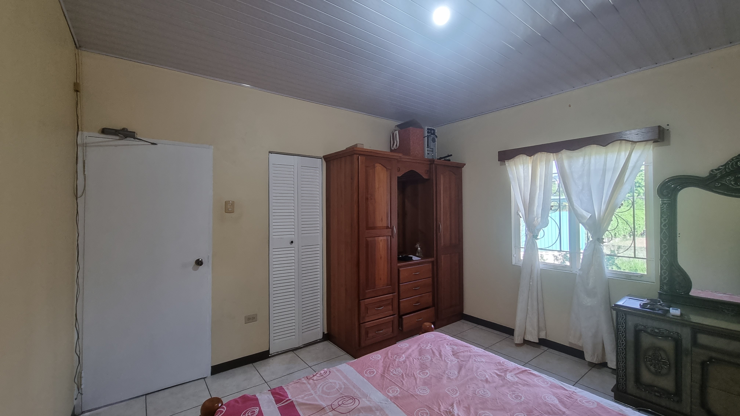 Bedroom in a fully furnished house for sale, featuring a large wooden wardrobe, a white closet door, and a bed with a pink bedspread.