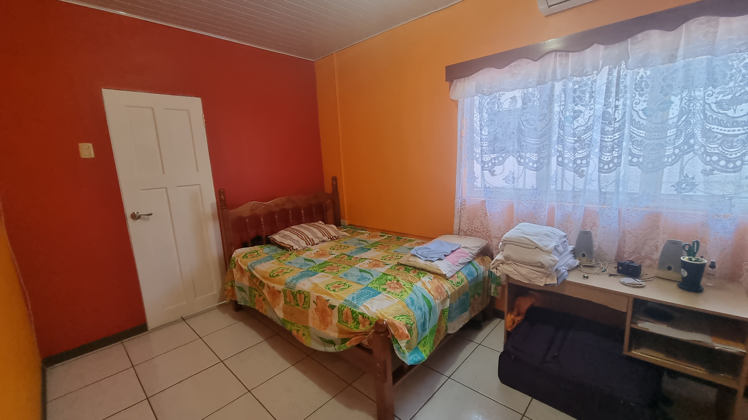 A colorful third bedroom in a fully furnished house for sale, with a wooden bed, red and orange walls, a desk, and an air conditioning unit.