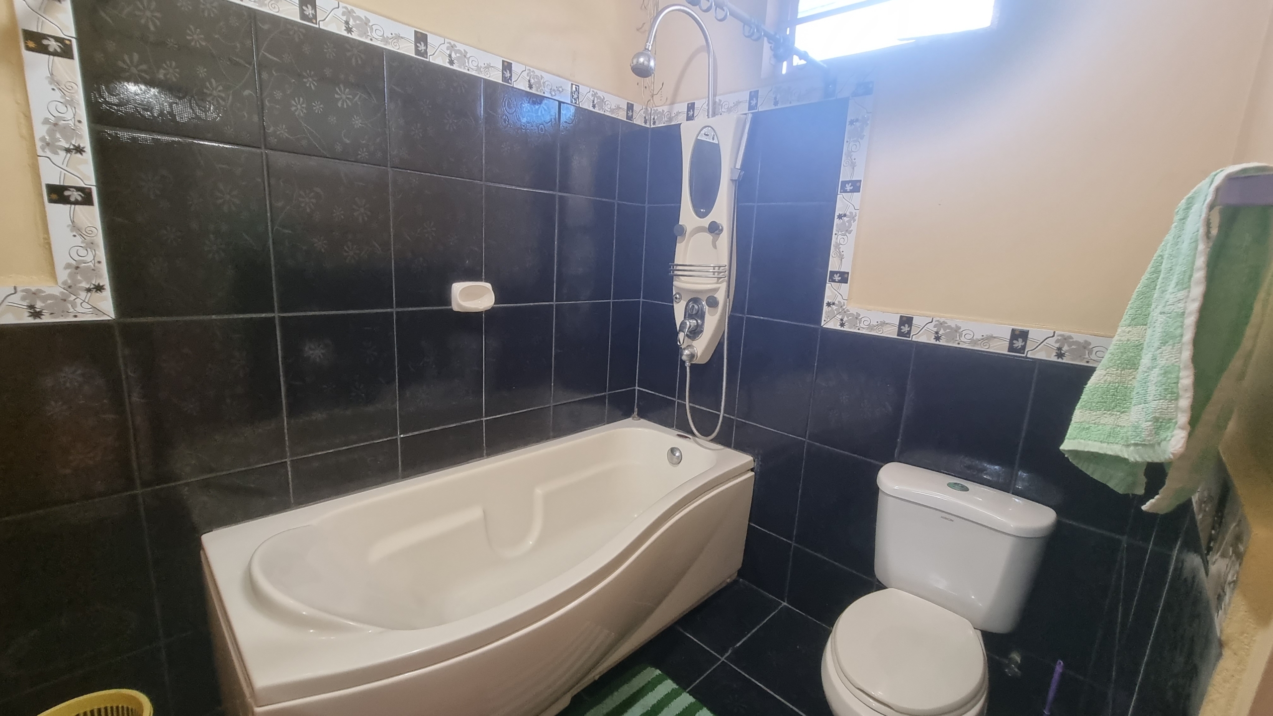 A modern bathroom in a fully furnished house for sale, featuring dark tiled walls, a white bathtub, toilet, and shower fixture.