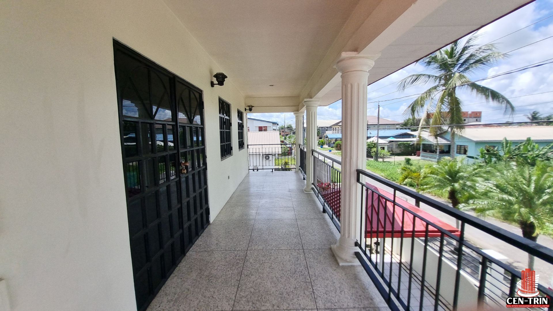Panoramic view from the upper balcony of an apartment building for sale in Chaguanas, showing tiled flooring, white pillars, black railings, and the surrounding neighborhood, highlighting rental investment potential.