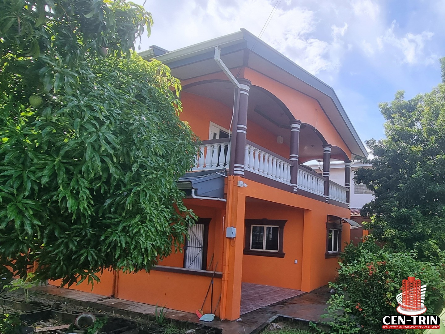 A vibrant orange, two-storey fully furnished house for sale in Balmain Gardens, Couva, with a white balcony railing, dark brown pillars, and lush green trees in the foreground.