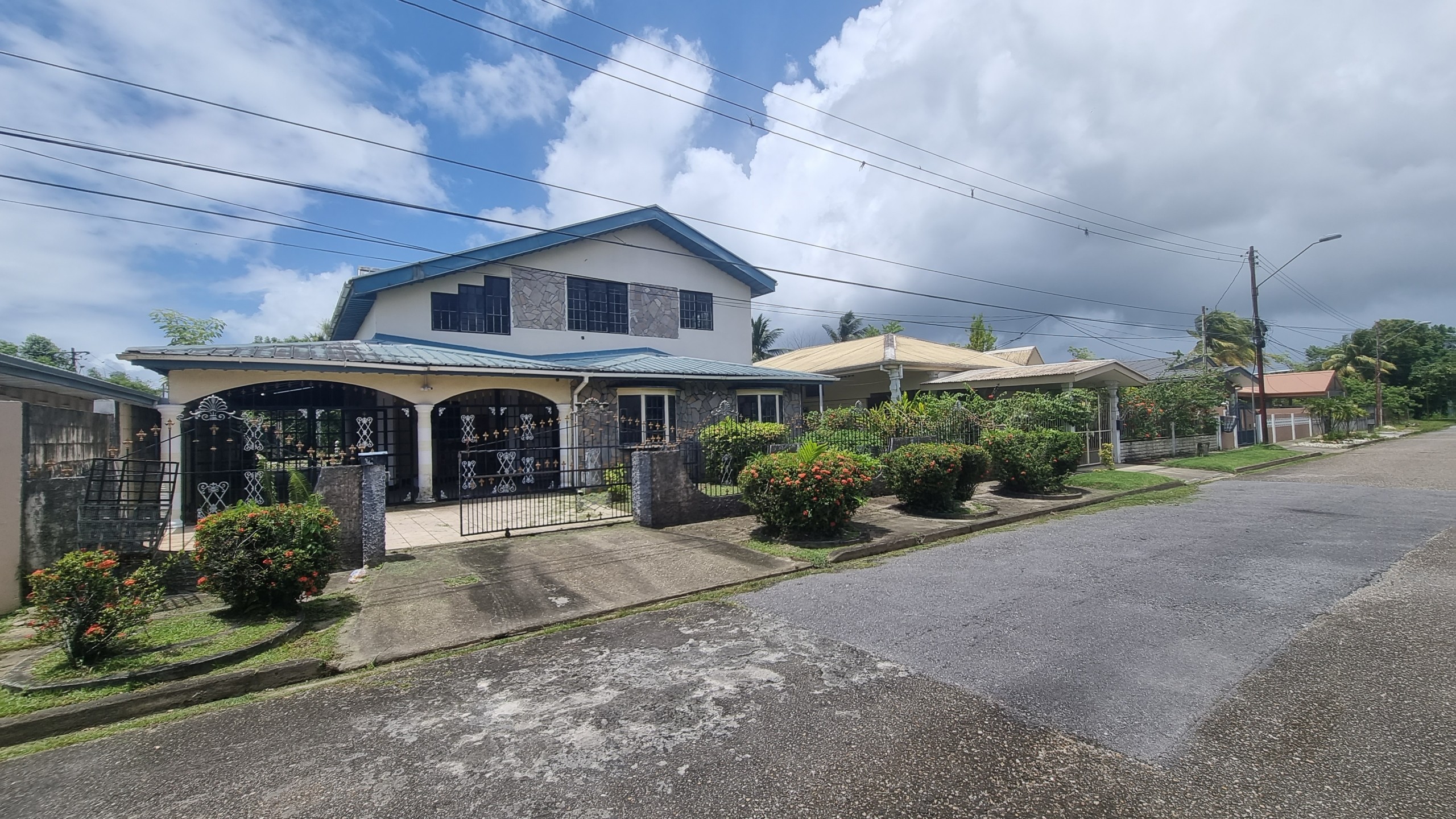 A two-story residential property for sale in Santa Rosa with a light-colored exterior and a blue roof. The first floor features stone-like siding and windows with protective bars. The front of the house has a black wrought-iron gate and a driveway leading to a covered carport, with green shrubs and plants in the front yard.