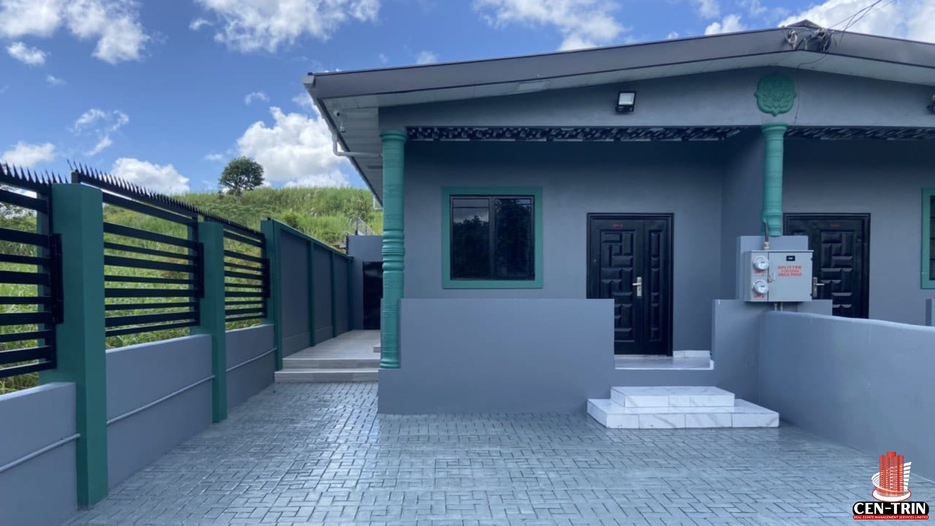A modern, two-year-old Barrackpore Duplex for Sale featuring a grey exterior with green trim and a paved driveway. The property is secured by a black metal fence with razor wire and is part of a Smart Home Trinidad security system, representing a prime Trinidad Investment Property suitable for Trinidad Airbnb Property revenue. Note the paved parking area and clear, well-maintained entrance.
